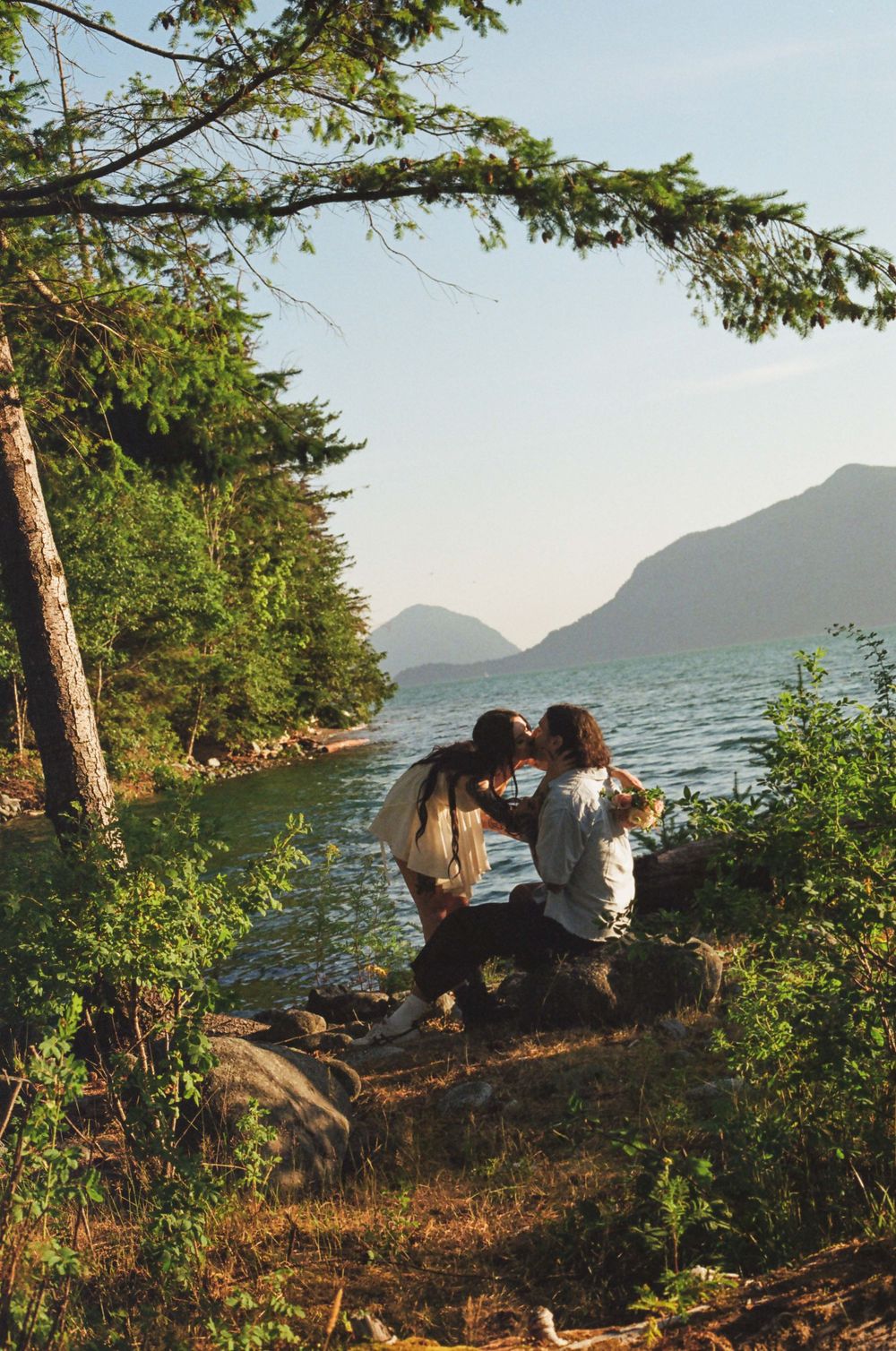 Two people relaxing on rocky outcrop overlooking serene mountain lake at sunset.