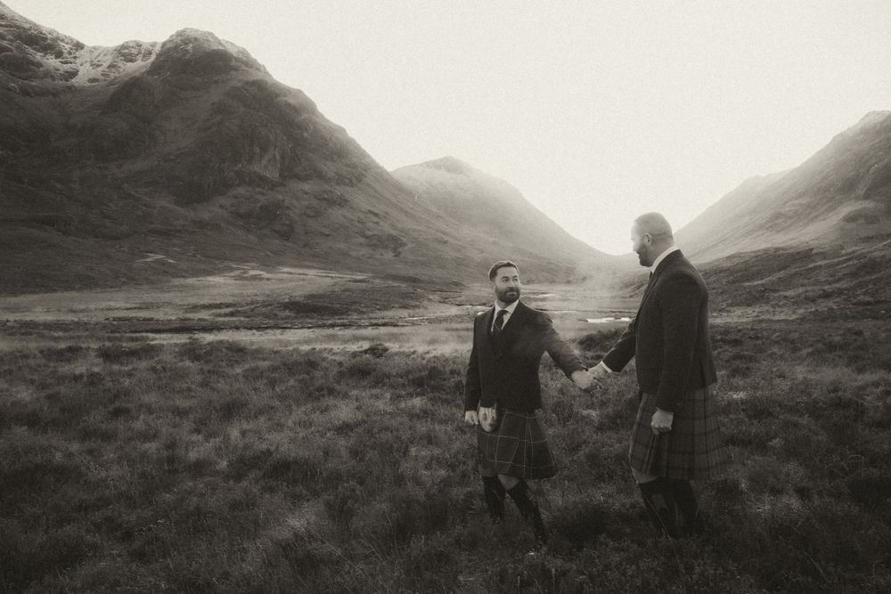 Black and white photograph of two people in traditional Scottish kilts standing in a misty Highland valley landscape.