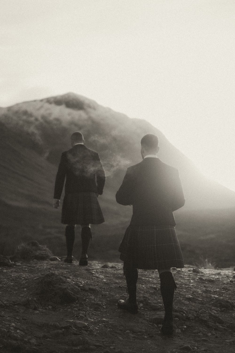 Two grooms in traditional kilts stand on a misty mountainside in Glencoe Scotland, photographed from behind in black and white.
