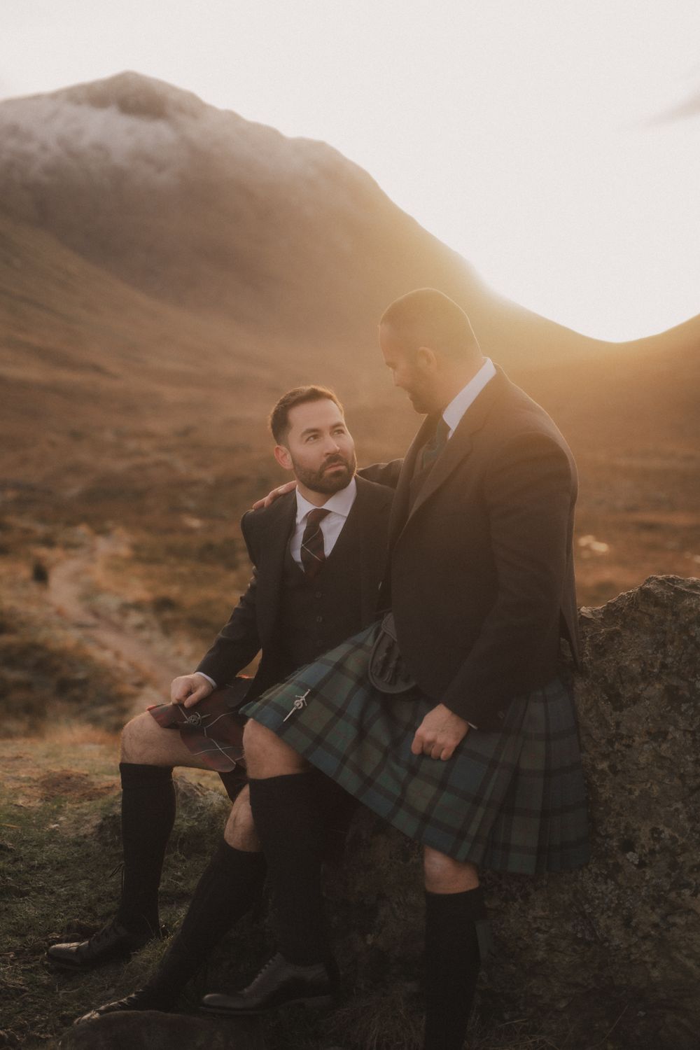 Two people in kilts sit together on rocks at sunset in a mountainous Scottish landscape.