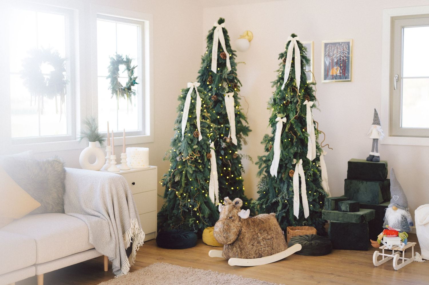 Modern living room decorated with tall evergreen Christmas trees, a rocking horse, and white winter wreath decor.