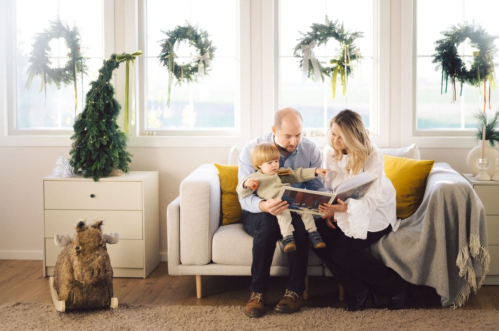 A cozy living room scene with white wreaths on the wall, a Christmas tree, and a family reading together on a white sofa.