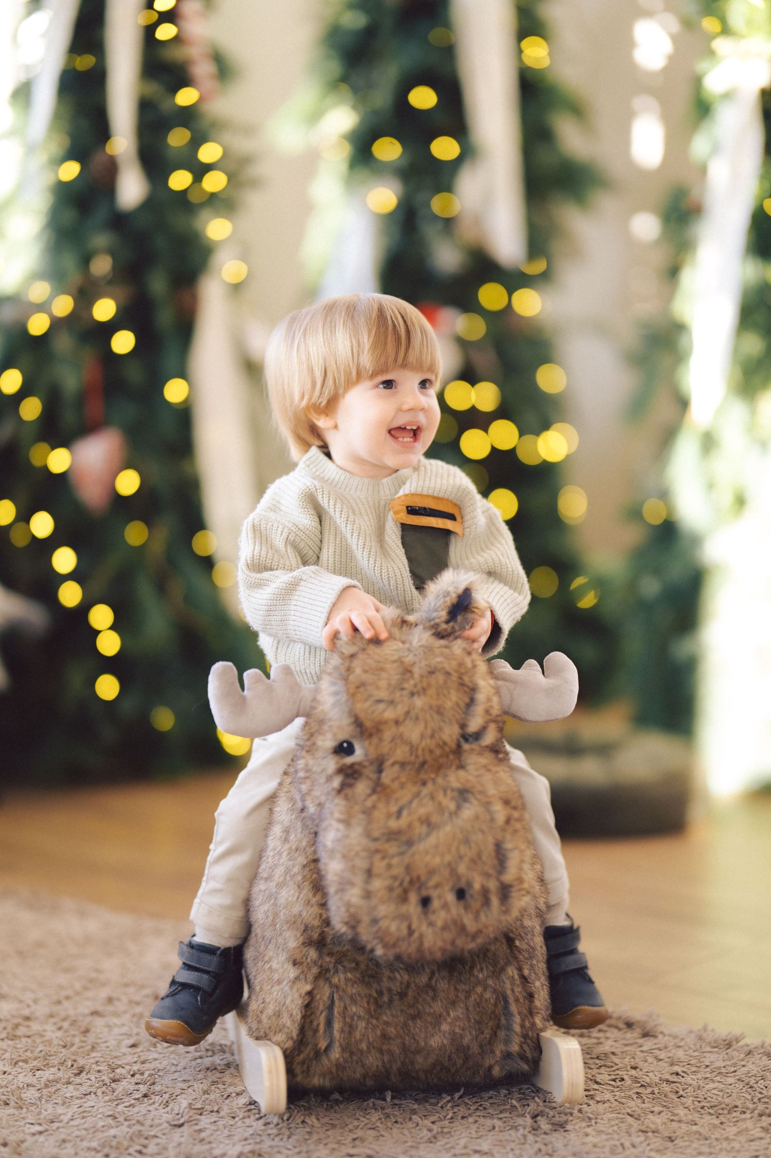 A young child in a white sweater rides a plush reindeer rocker against a background of Christmas lights.