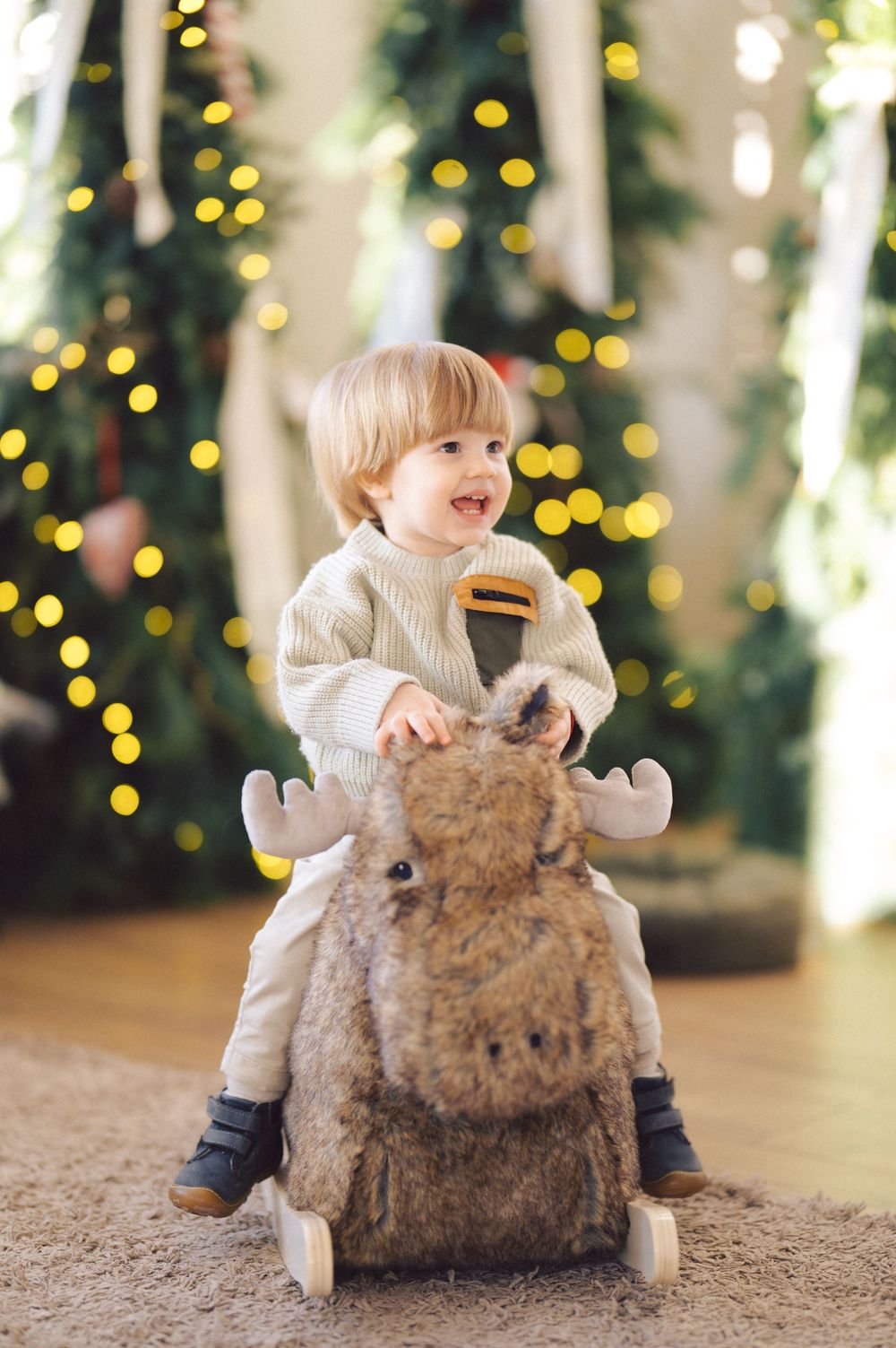 A young child in a white sweater rides a plush reindeer rocker against a background of Christmas lights.