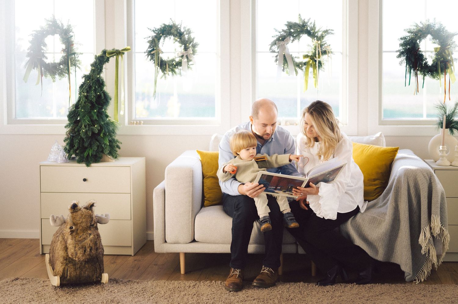 A cozy living room scene with white wreaths on the wall, a Christmas tree, and a family reading together on a white sofa.