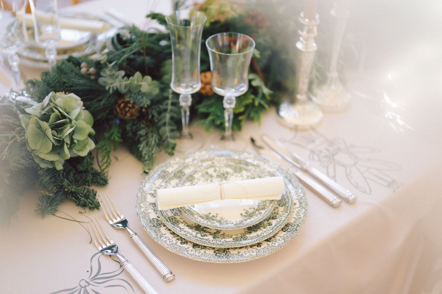 Elegant holiday table setting with floral china plates, crystal glasses and greenery centerpiece on white linen.