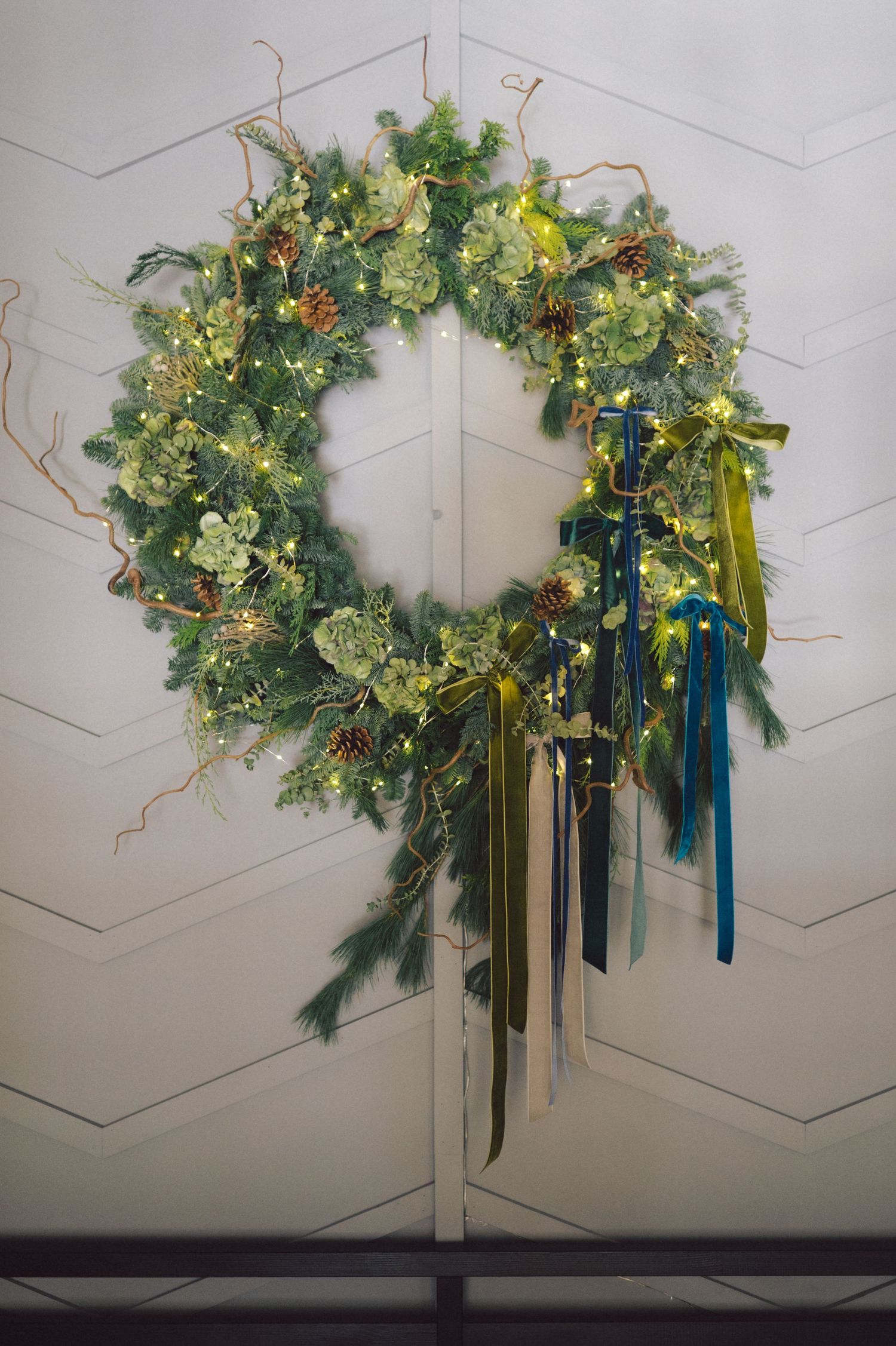 A festive Christmas wreath adorned with greenery, gold accents, and cascading blue and yellow ribbons hangs on a white wall.