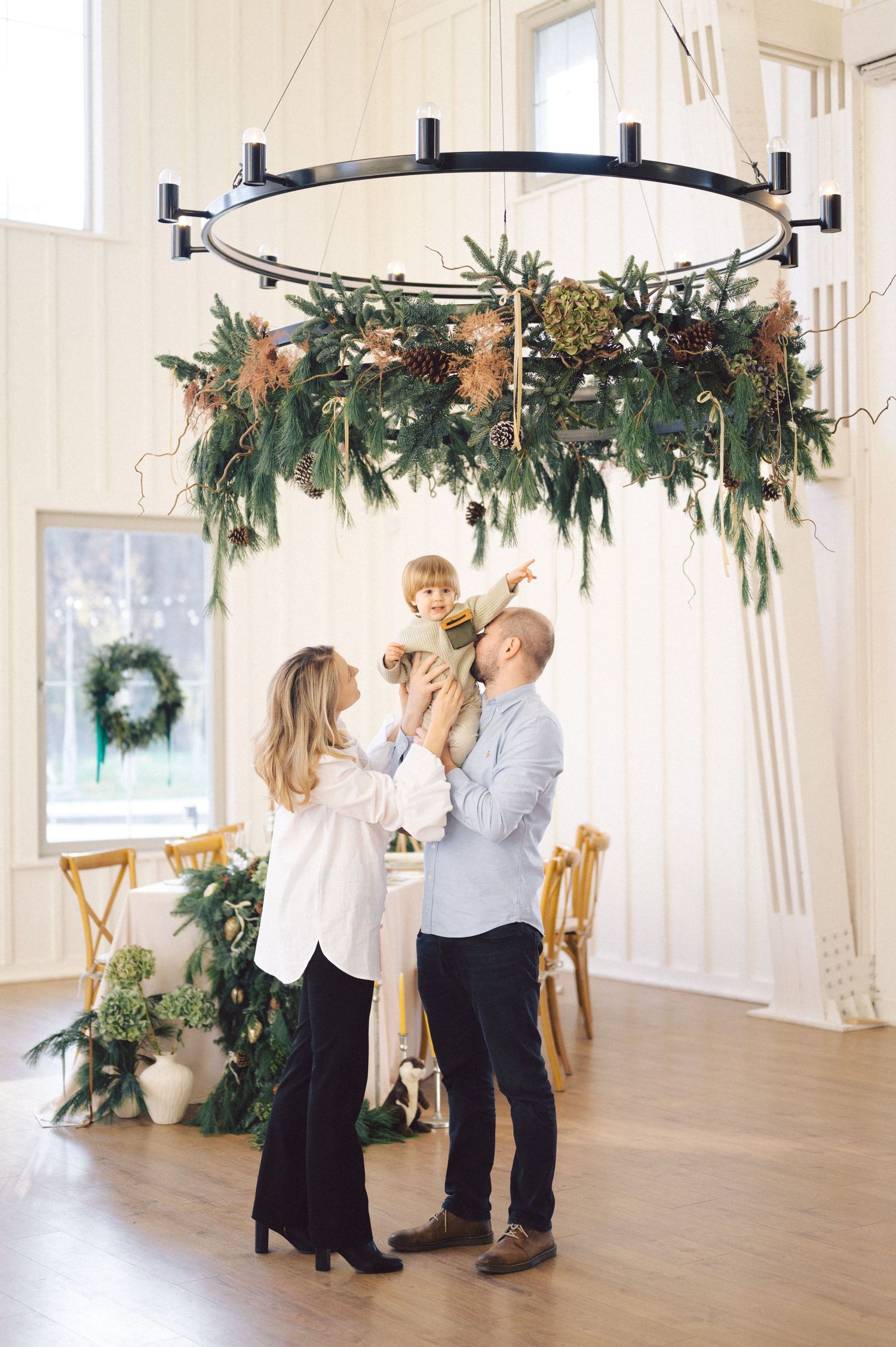 Family gathering beneath decorative chandelier adorned with evergreen foliage and holiday greenery in bright interior space.