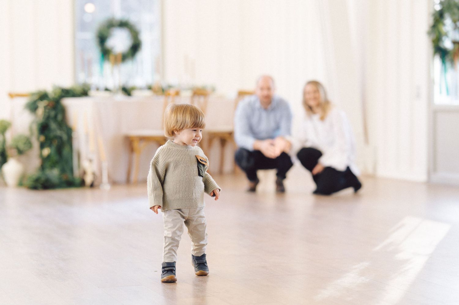 A young child in a gray outfit takes early steps across a bright white room with adults watching in the background.