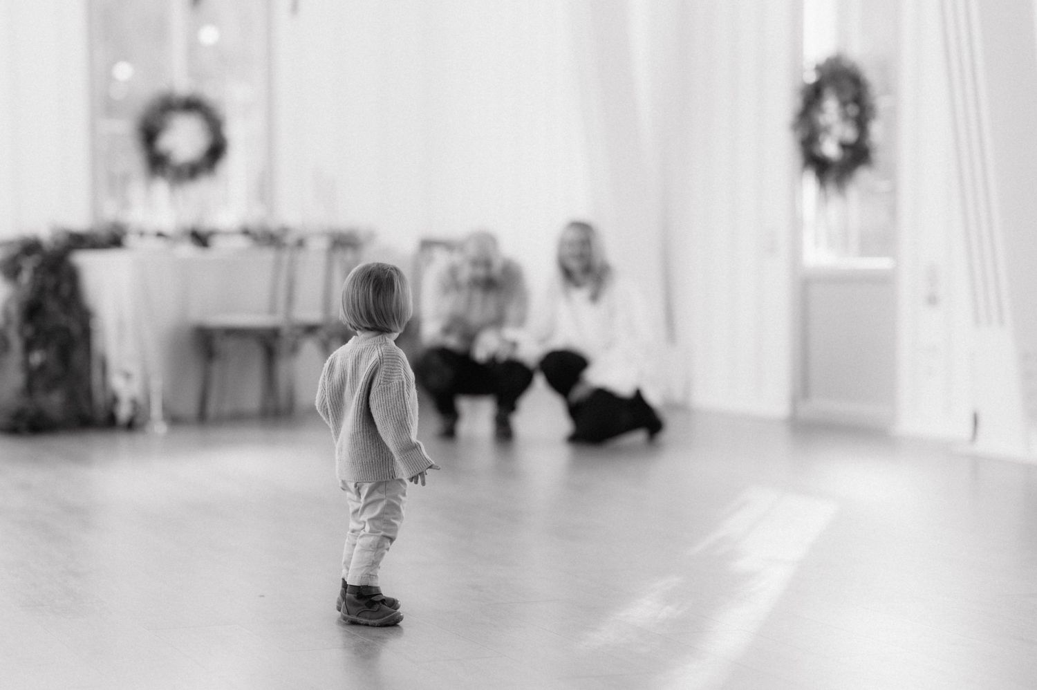 Small child stands alone in a minimalist white room with Christmas wreaths hanging on the walls in black and white.