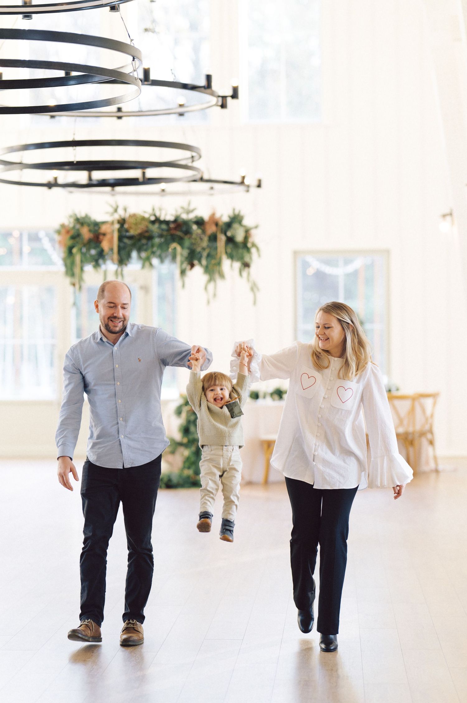 In a bright white studio with chandeliers overhead, two people walk and laugh together in coordinated outfits.