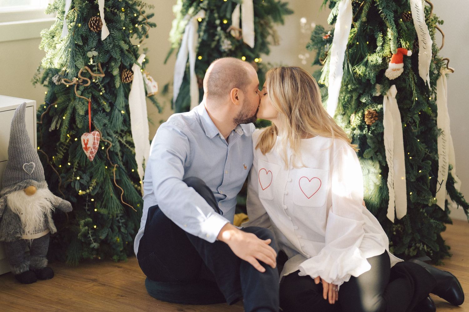 Couple sitting together by decorated Christmas tree surrounded by white drapes and holiday decor.