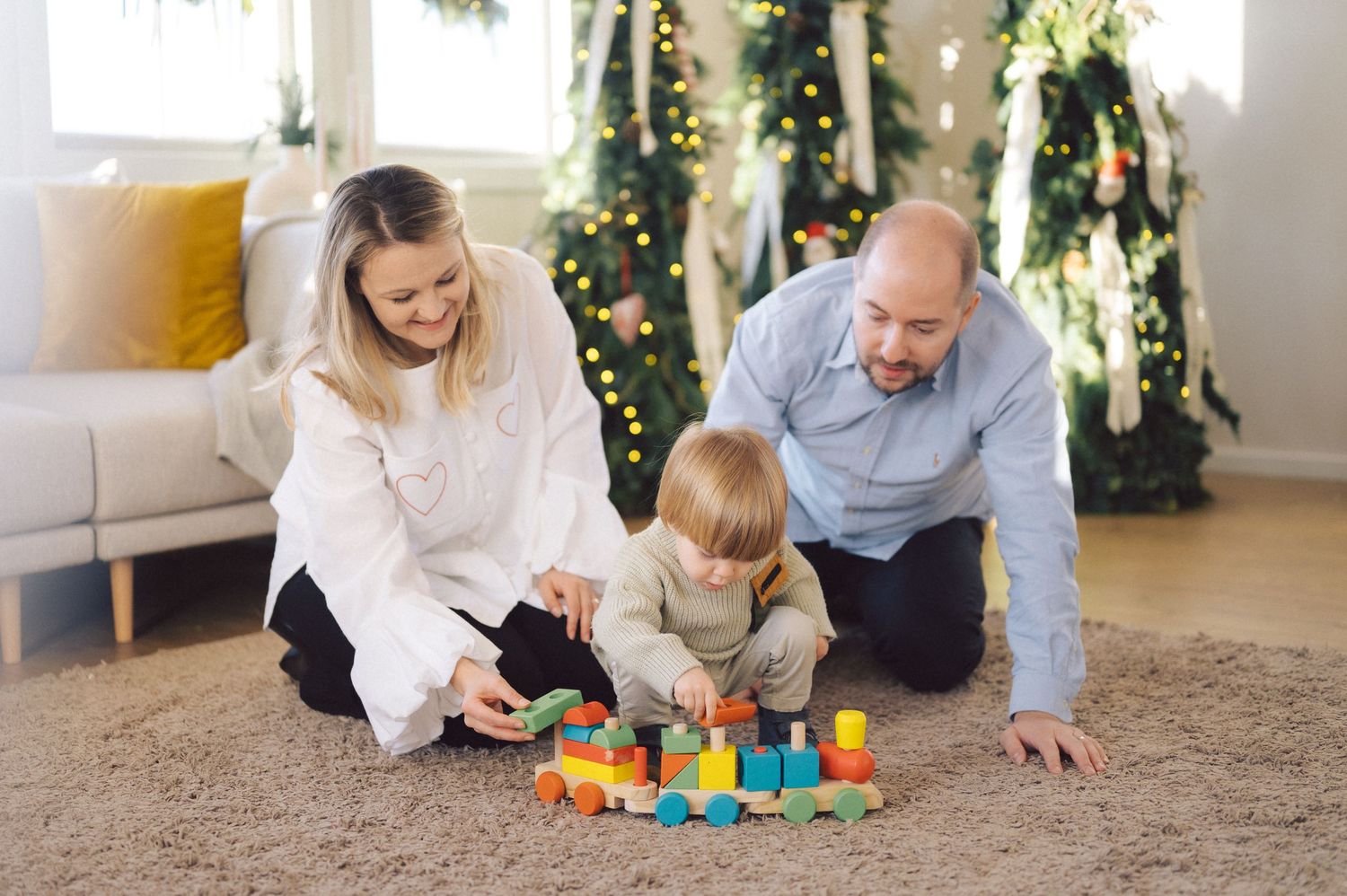 Family sitting on carpeted floor playing with colorful toy blocks near Christmas tree.