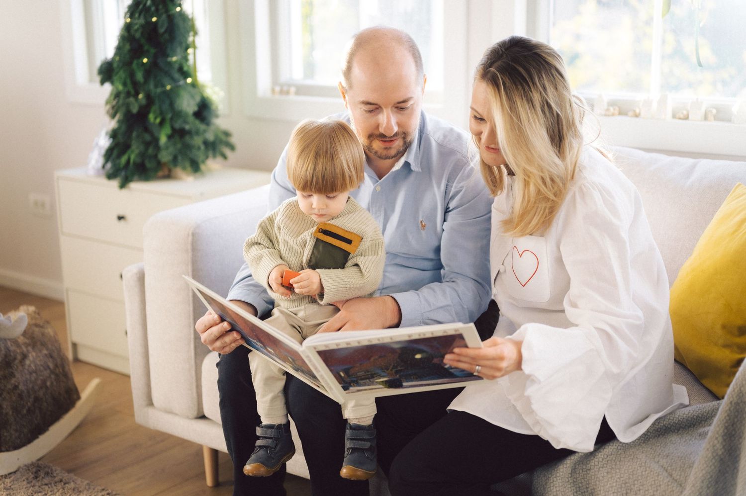 Family reading a book together on couch in bright living room with Christmas tree in background.