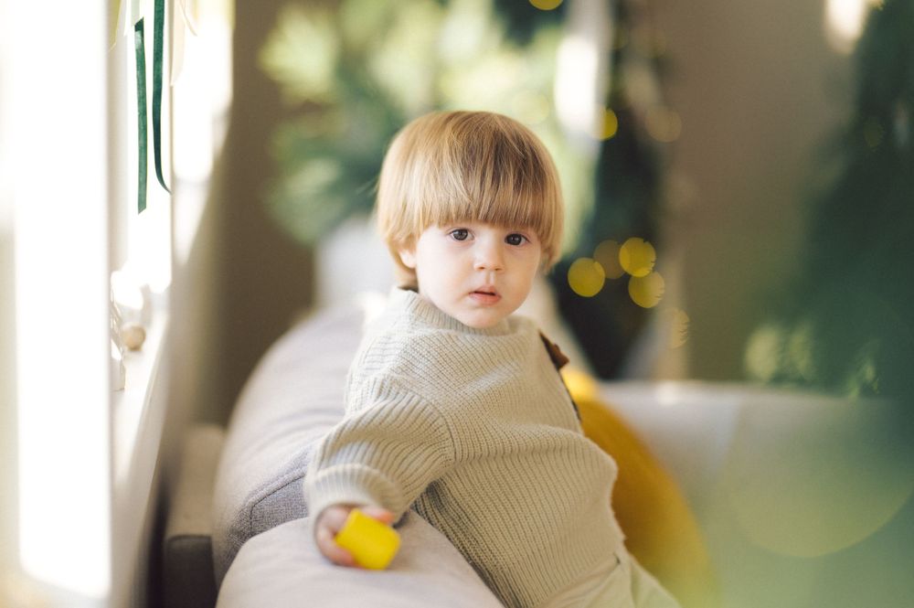 Small child in cozy beige sweater holding yellow toy sits near window with soft bokeh lights in background.