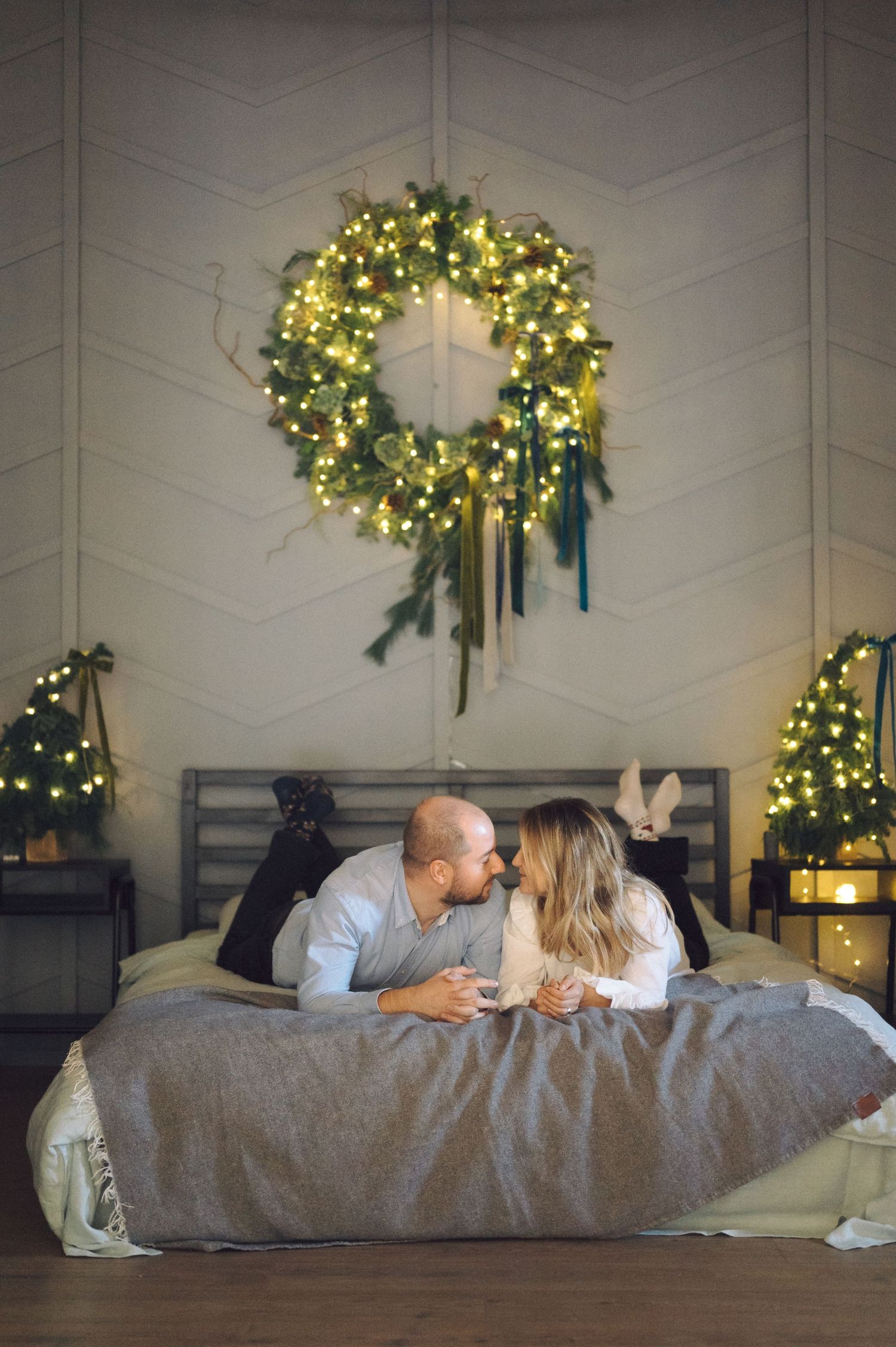 A couple lies on a gray bed beneath a glowing Christmas wreath and holiday decorations in a cozy bedroom setting.