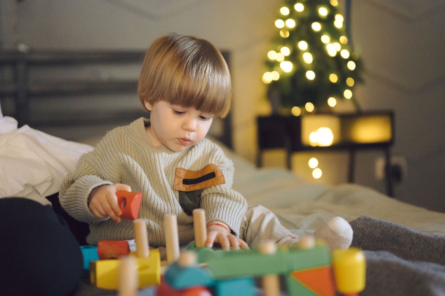 Small child plays with colorful building blocks and toys near a lit Christmas tree in a cozy living room.