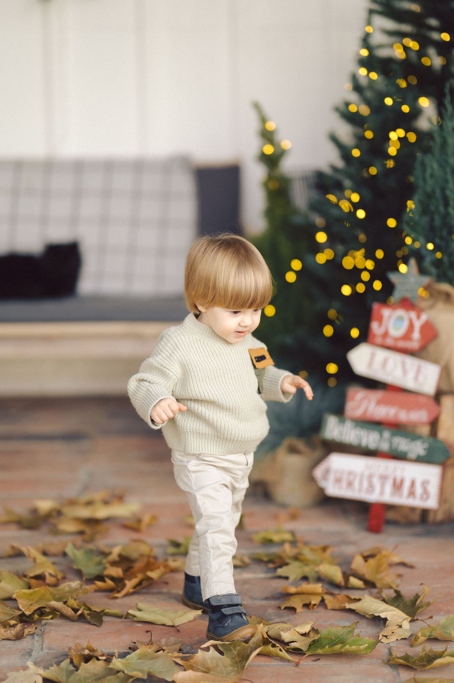 Small child in cream sweater plays with autumn leaves in front of a decorated Christmas tree and holiday signage.