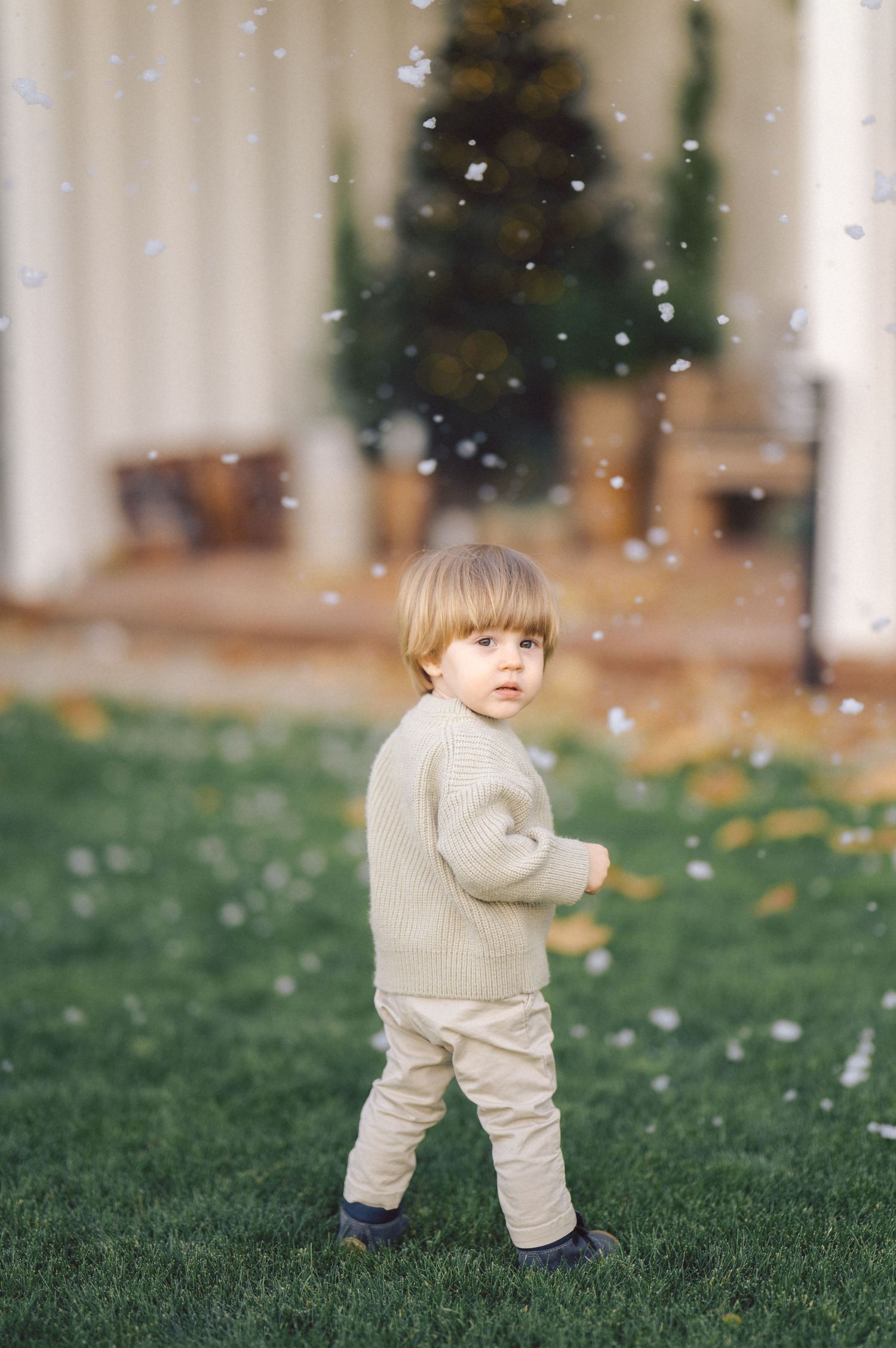 A small child wearing a beige sweater stands on grass while autumn leaves fall in the peaceful outdoor setting.