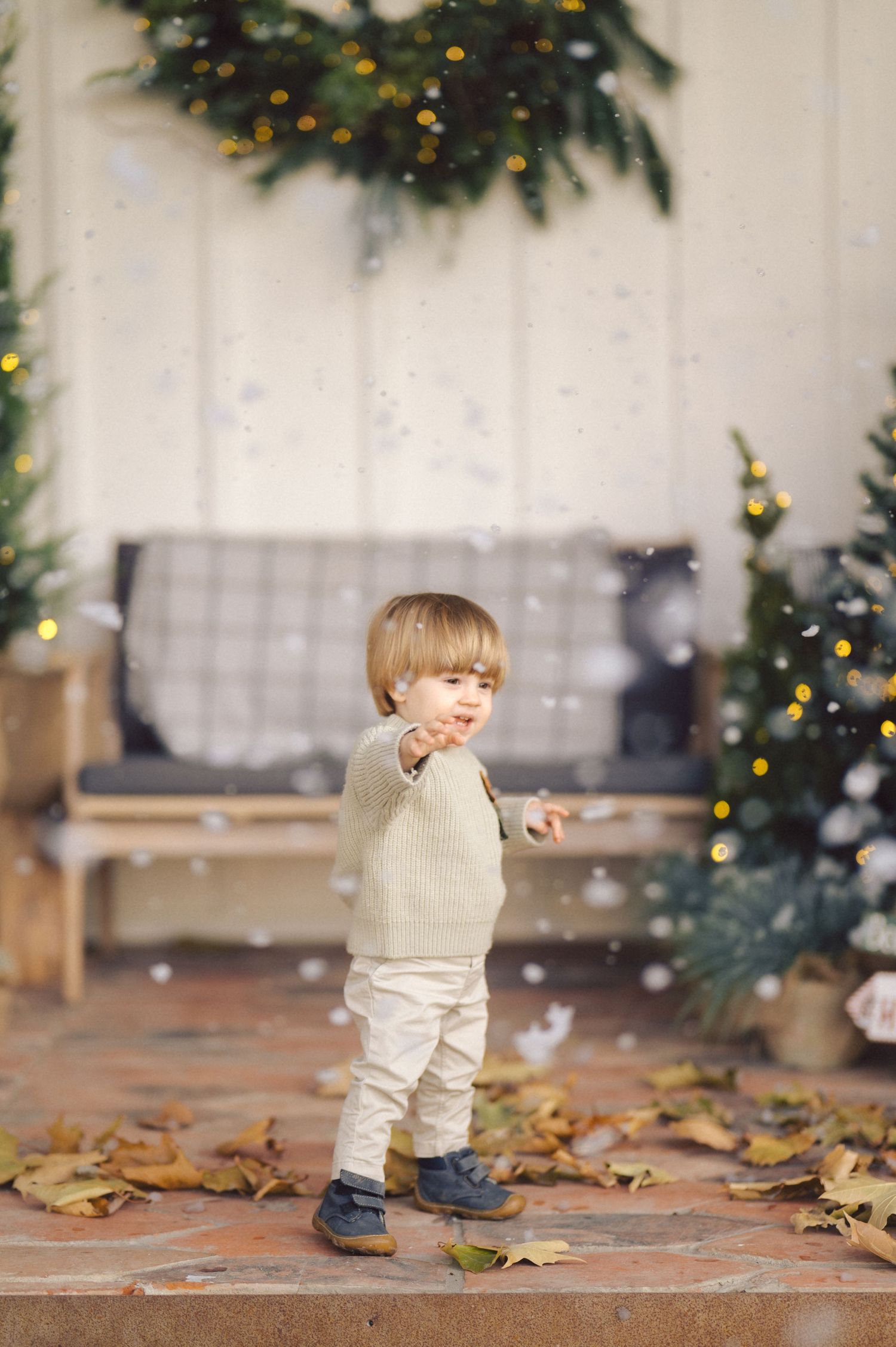 Small child in beige sweater and light pants plays with falling leaves in front of a Christmas decorated room.