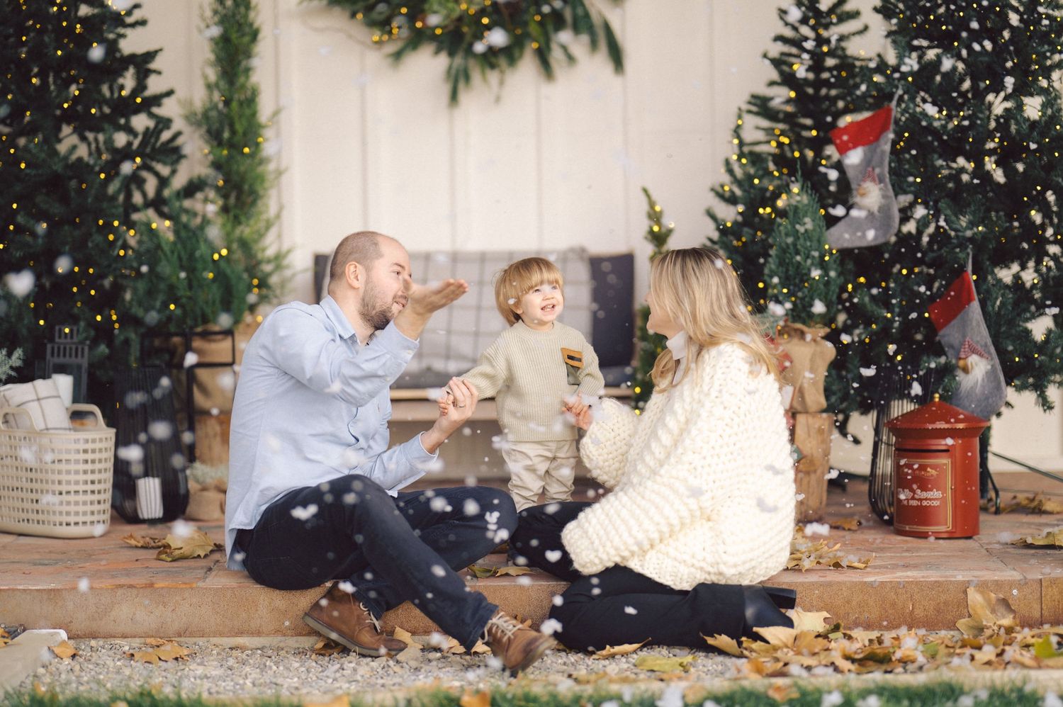 Family sits together amid festive Christmas decorations while snow-like confetti falls around them in a cozy indoor setting.