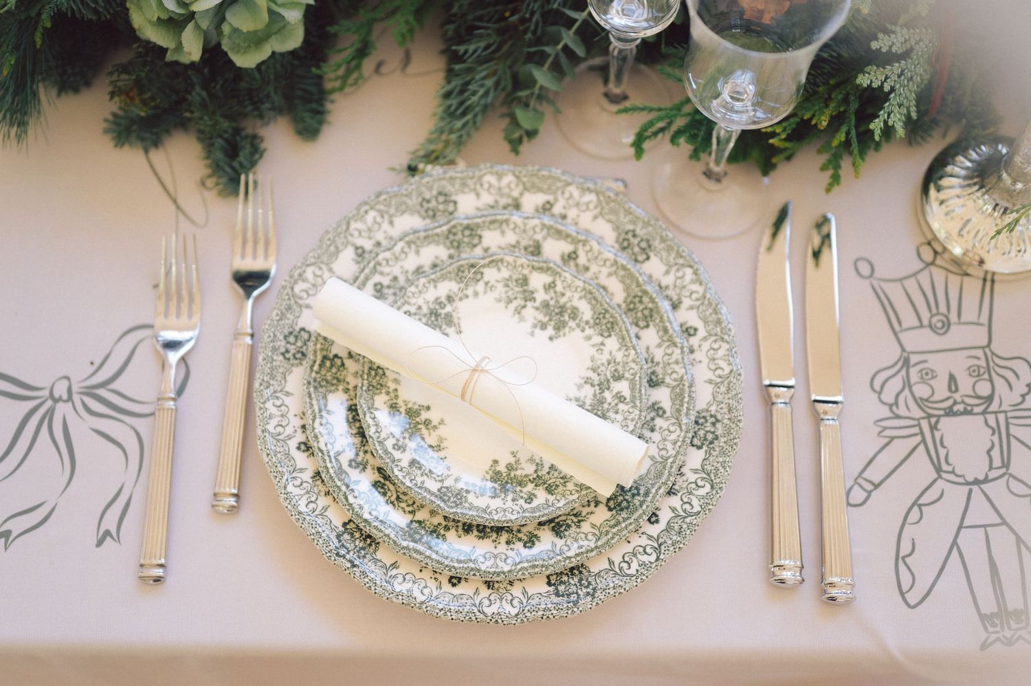 Elegant place setting with floral patterned china plates and silver cutlery arranged under holiday greenery decorations.