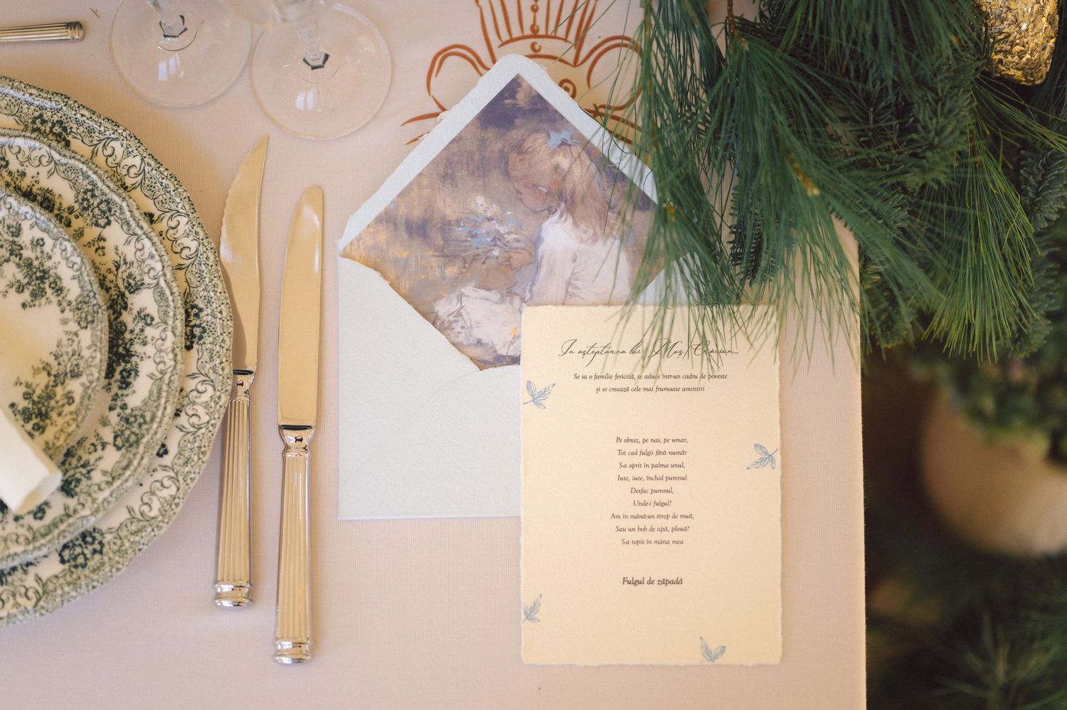 Wedding invitation and envelope displayed on a white table with evergreen branches and vintage china plate decor.