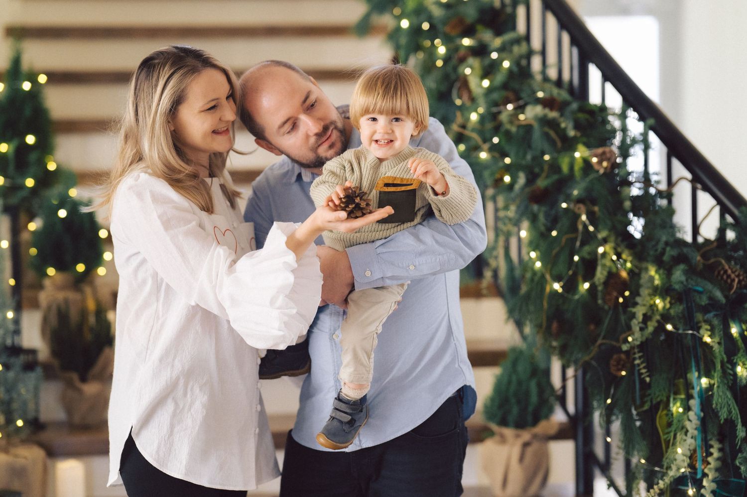 A couple poses with a baby for holiday photos in front of a decorated staircase with greenery and lights.
