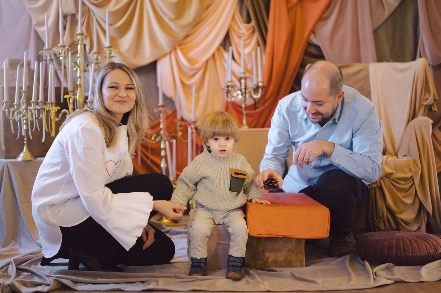 Family sits together in an elegant setting with draped fabric and chandeliers during a special occasion photoshoot.