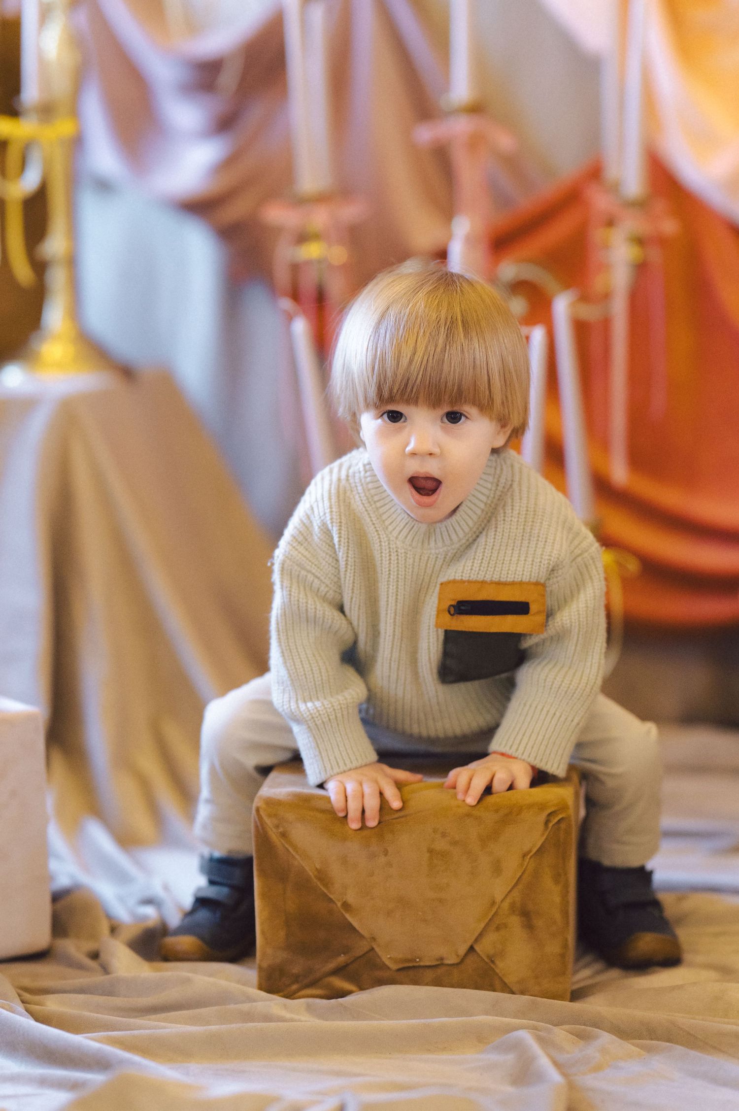 A small child in a beige sweater sits on a brown box against a draped fabric background.