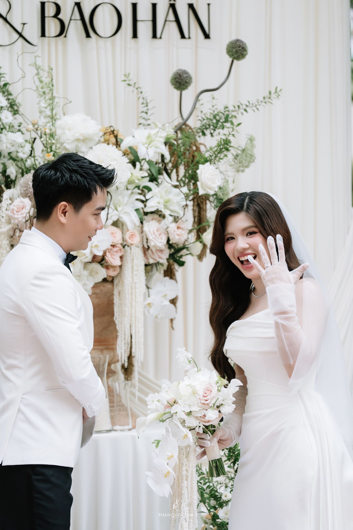 Bride shows off wedding ring while smiling at groom in white tuxedo at elegant floral ceremony backdrop.