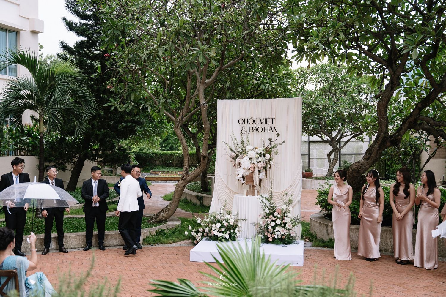 Outdoor wedding ceremony with guests holding clear umbrellas near a floral backdrop reading Quoc Viet and Bao Han.