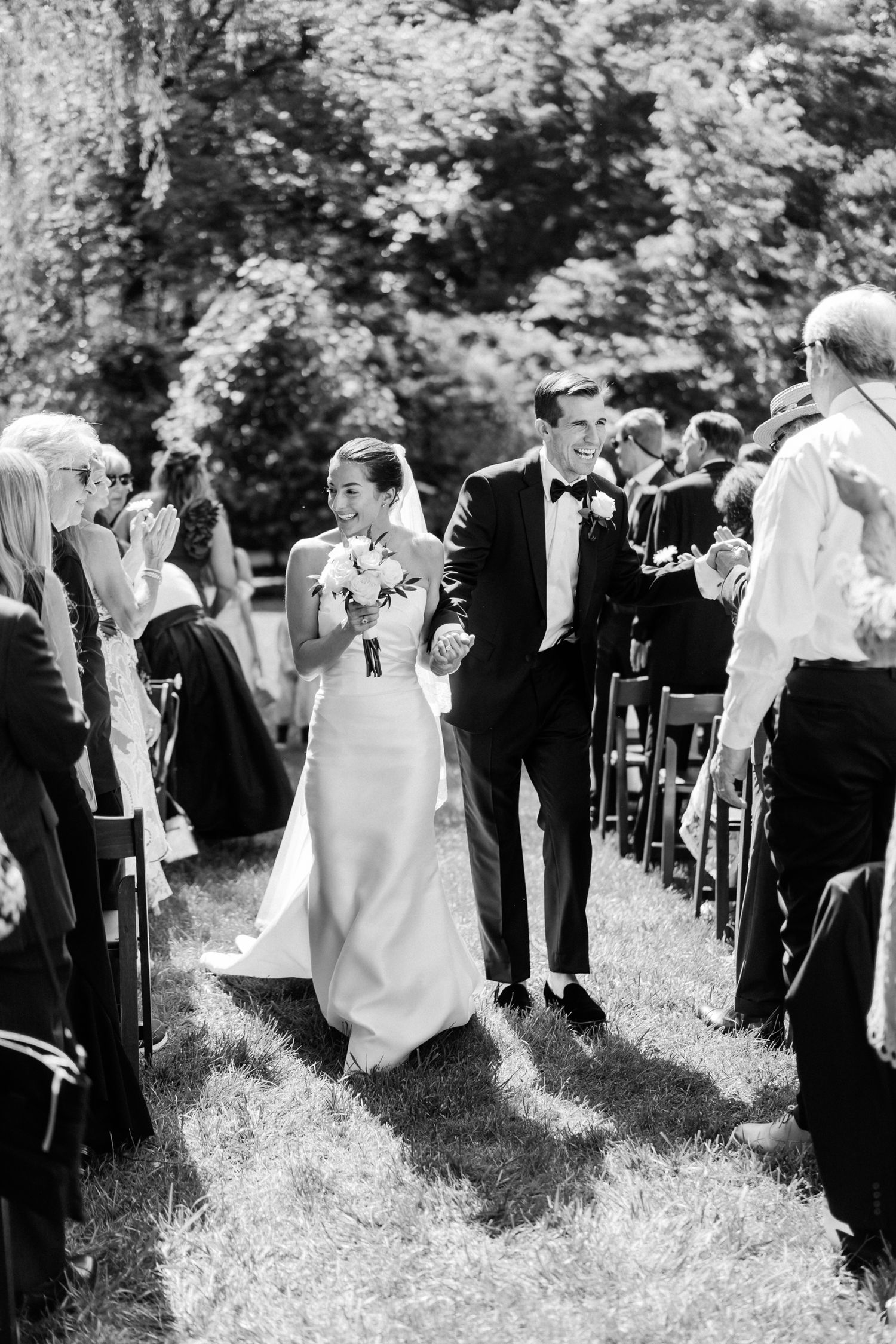 Black and white photo captures outdoor wedding processional with guests lining a grass path amidst trees.