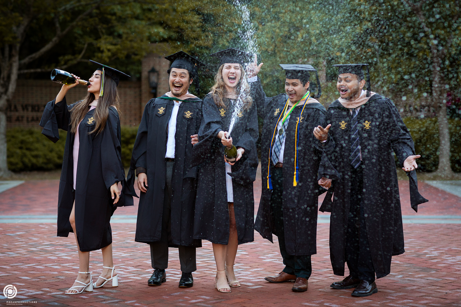 A group of graduates in black caps and gowns smile joyfully during commencement festivities with falling confetti.