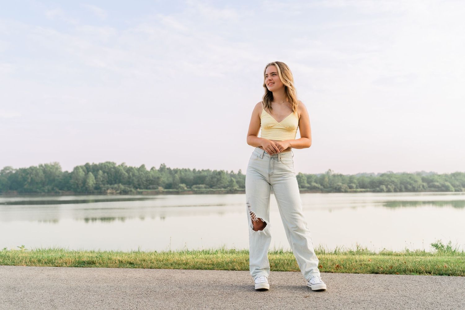 Senior in yellow crop top poses outdoors near a serene lake with trees in the background on the Maumee River in Perrysburg, Ohio.