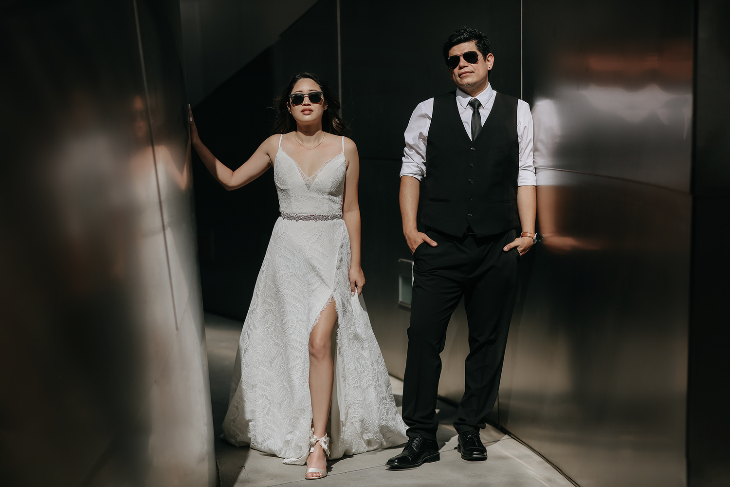 Stylish couple in formal attire poses in a moody elevator setting wearing sunglasses.