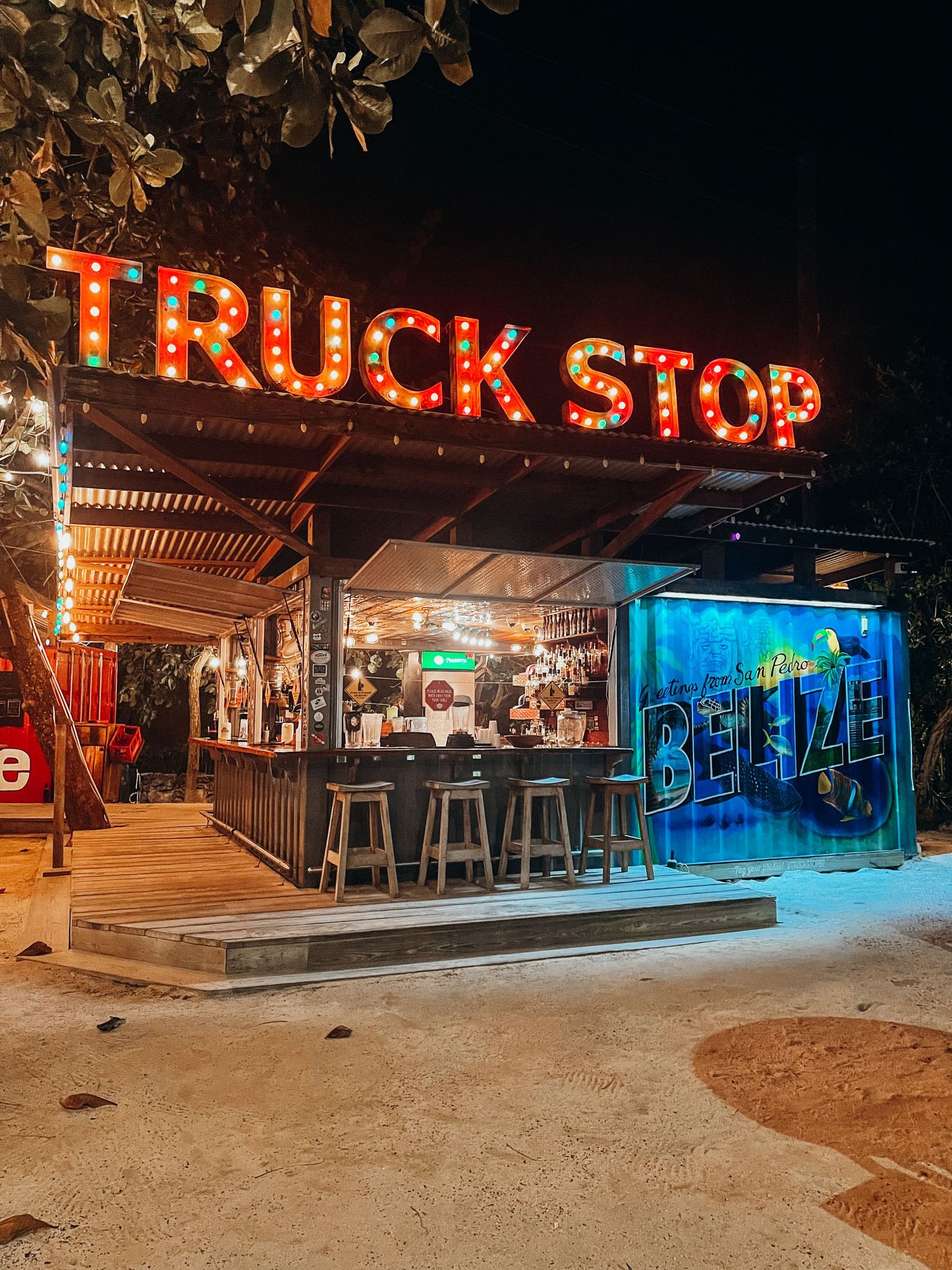 Night view of illuminated beach bar with neon 'Truck Stop' sign and outdoor seating area.