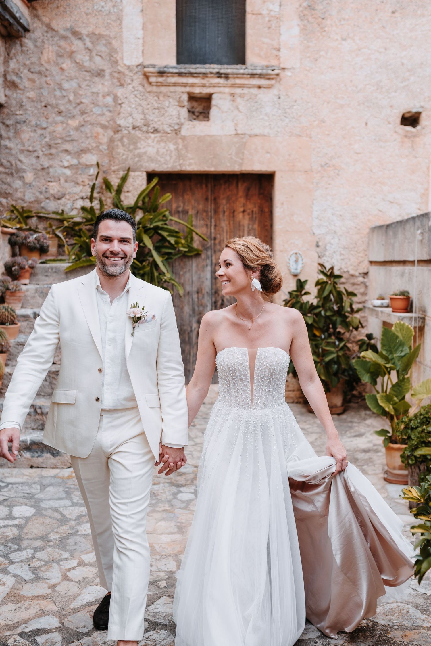 Ein Paar spaziert Hand in Hand in Hochzeitskleidung einen rustikalen Steinweg entlang, der in einem alten mallorquinischen Innenhof liegt.