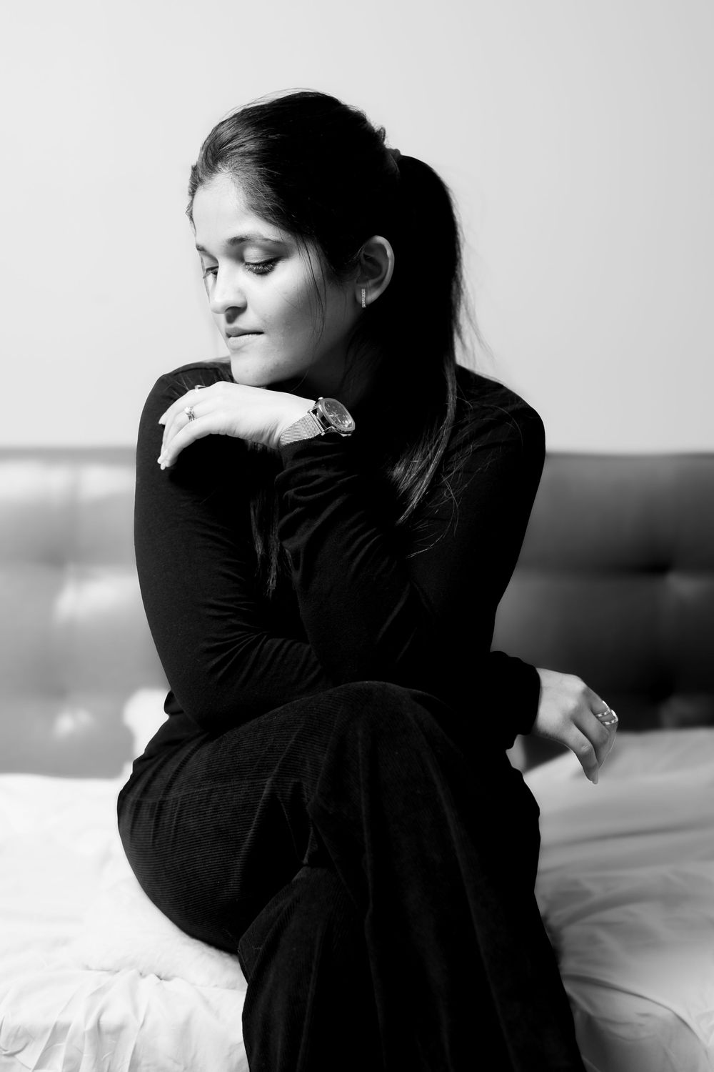 A black and white portrait of a figure in dark clothing sitting on a bed with a contemplative, downward gaze.