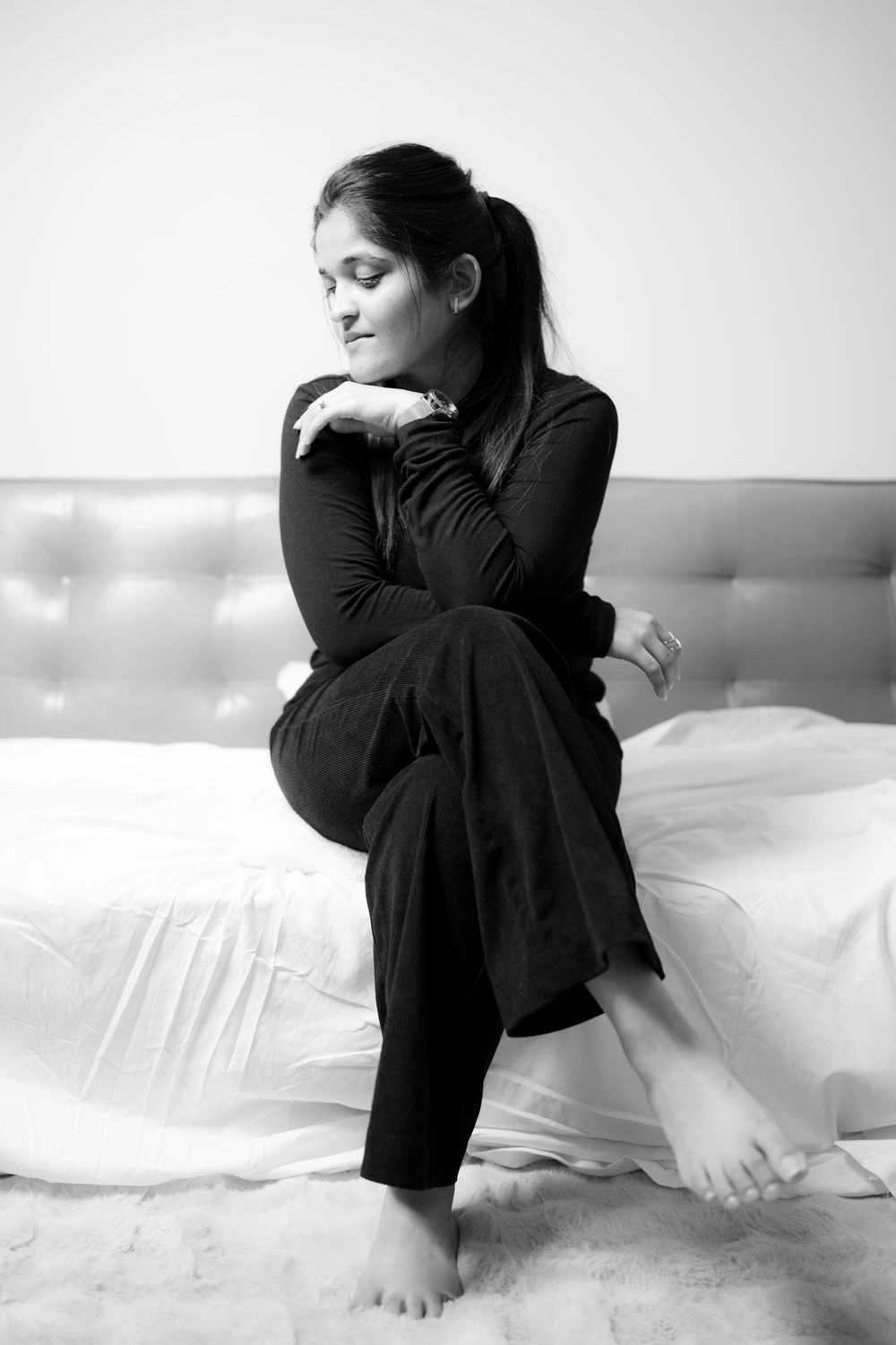 Black and white intimate portrait of person in dark clothing sitting on white sheets, with contemplative pose.