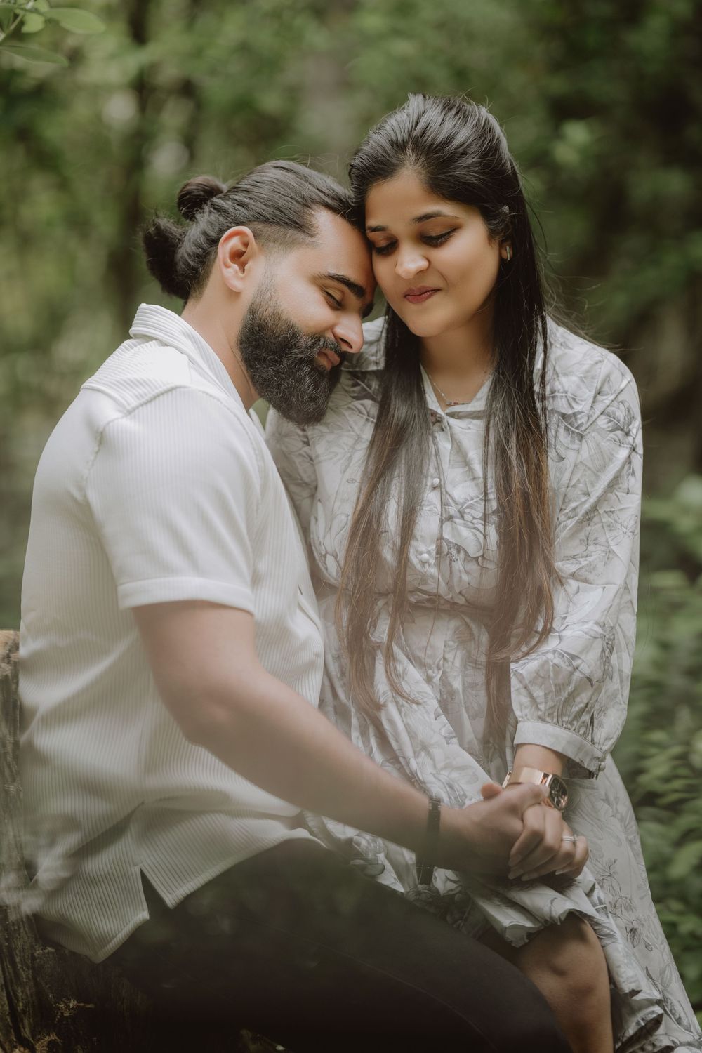 A couple shares a romantic moment outdoors in a forest setting, with her wearing a white dress and him in a white polo shirt.