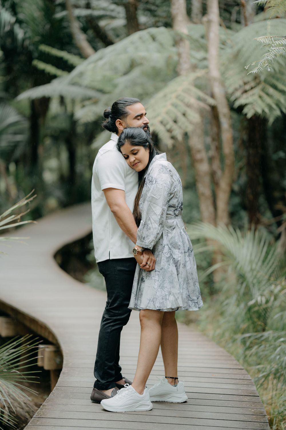A romantic couple shares tender moments on a wooden boardwalk surrounded by lush ferns and forest greenery.