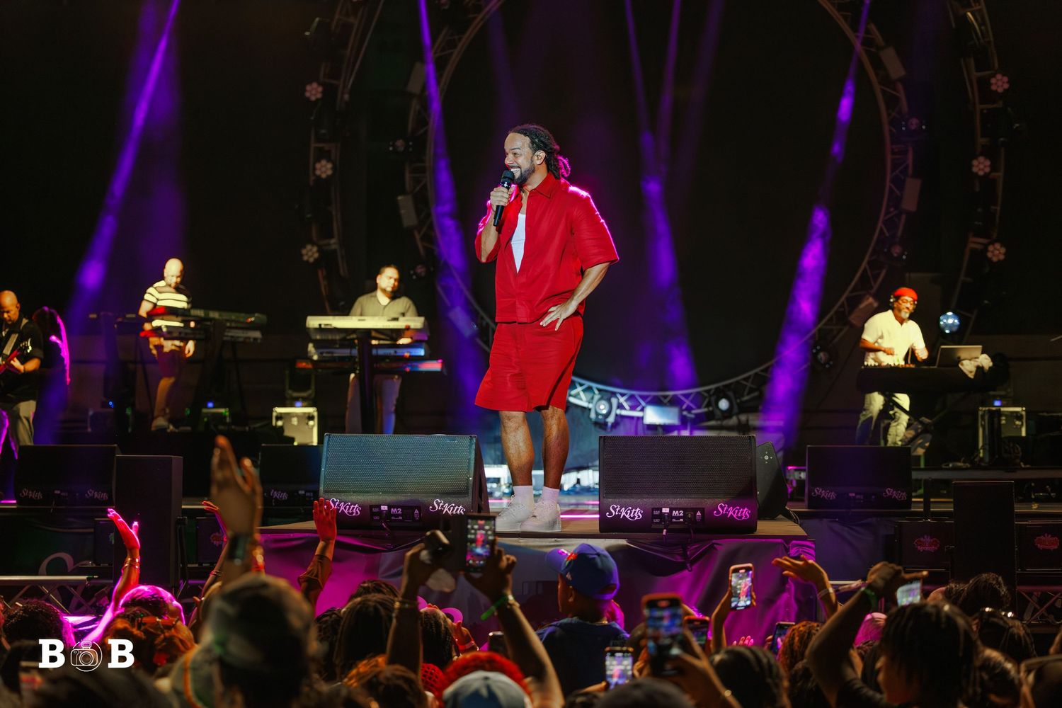 Kes is performing live at the St. Kitts Music Festival 2025, wearing a red outfit, as colorful stage lights illuminate the performance.
