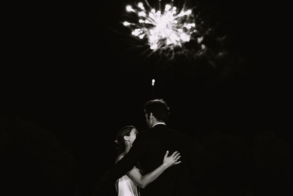 A couple embraces under a bursting firework in the night sky creating a romantic black and white moment.