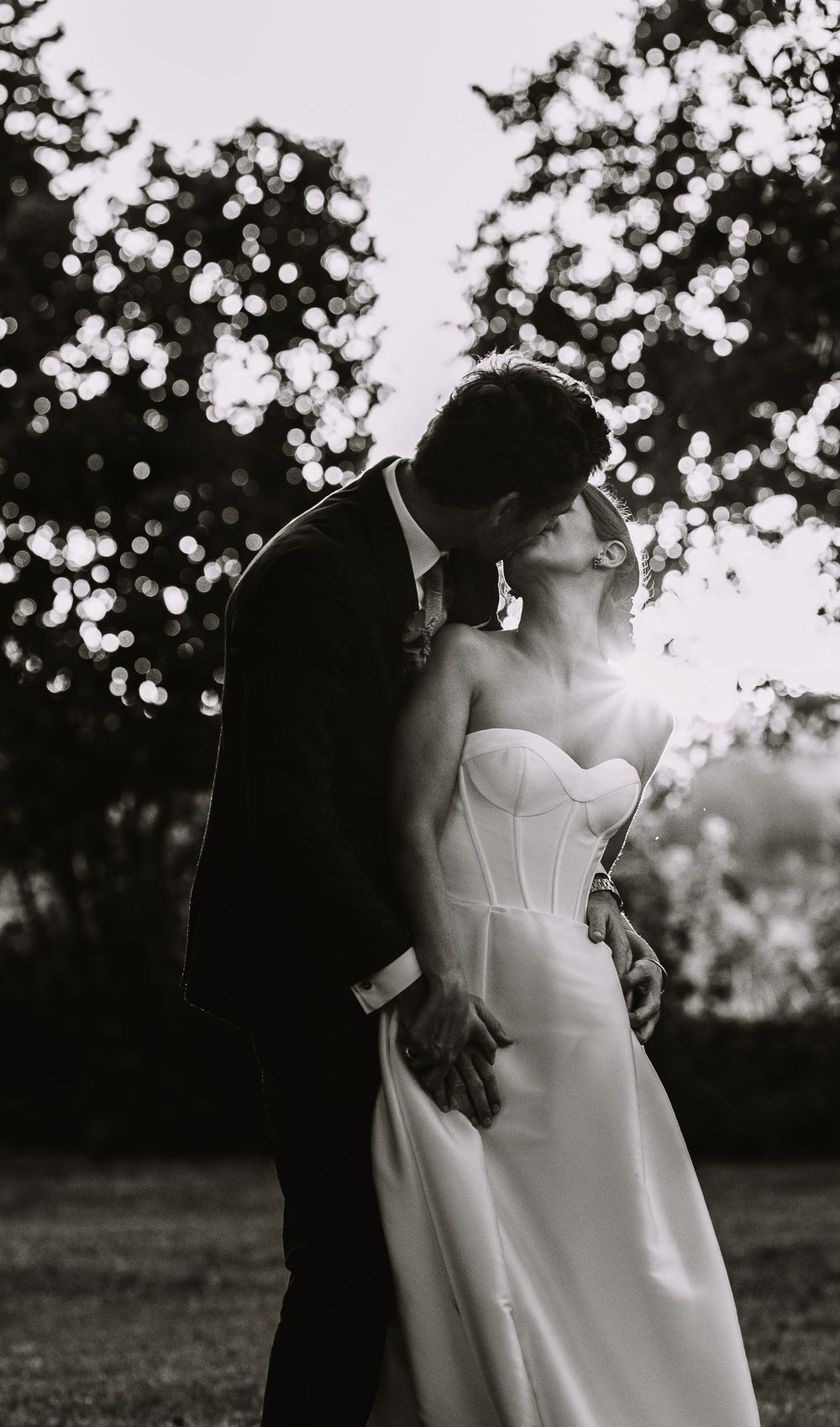 A romantic black and white wedding photo shows a couple in formal attire embracing outdoors under trees.