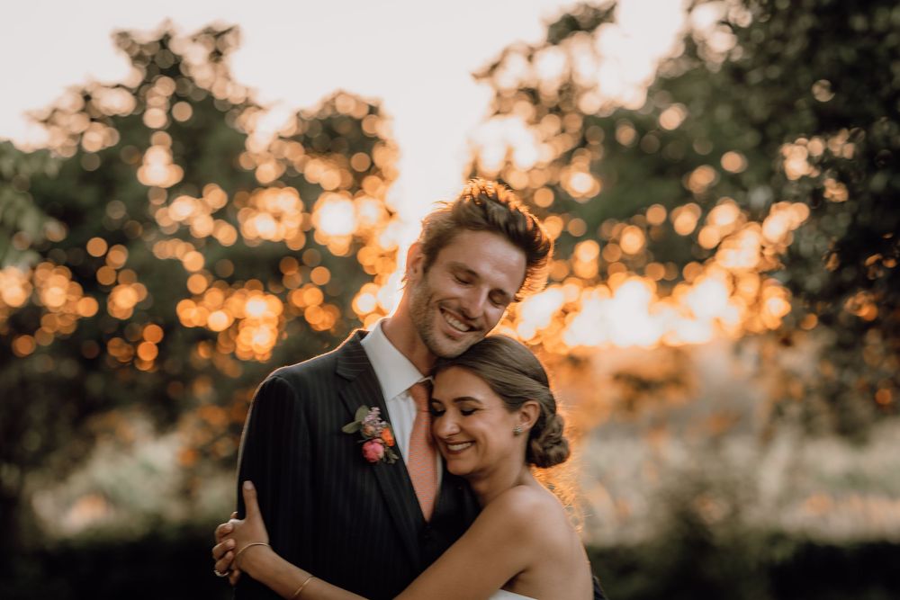 A happy couple embraces outdoors during golden hour sunset with bokeh lights and trees in the background.