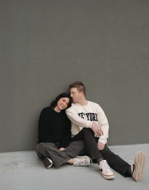 A couple in casual wear sits close together against a gray wall, with one wearing a New York sweatshirt.