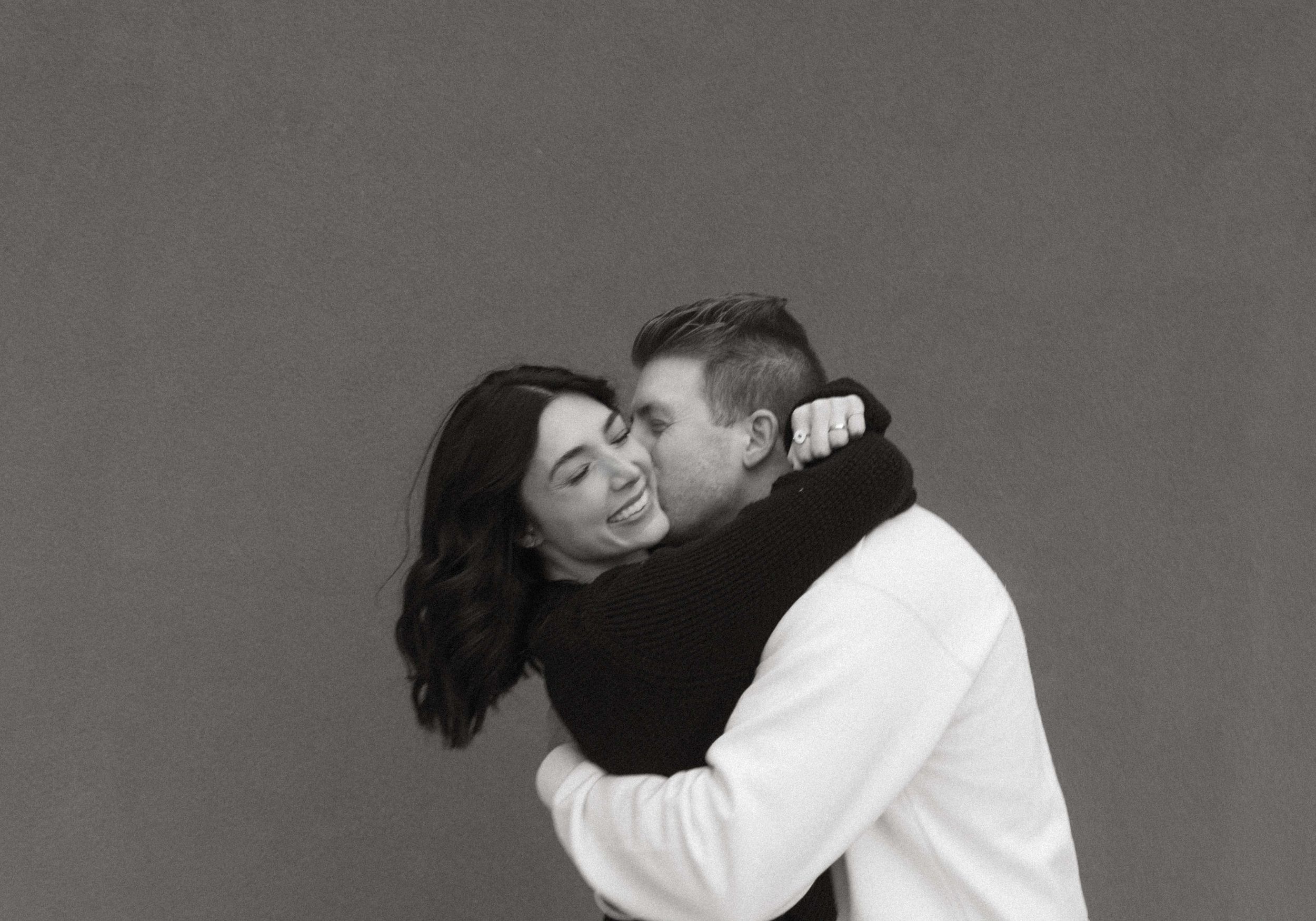 A couple embraces joyfully in a black and white portrait against a plain background.