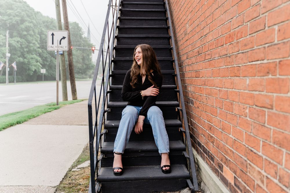 Candid fashion pose on outdoor metal staircase against brick wall wearing jeans and black top.
