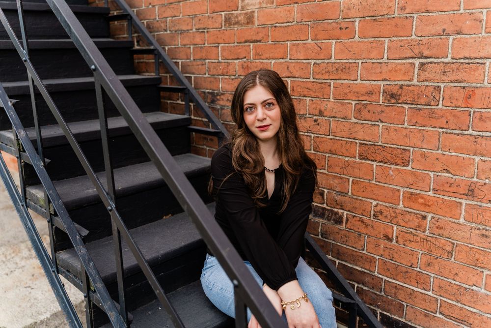 Relaxed pose against brick wall and metal stairs in casual denim and black outfit.
